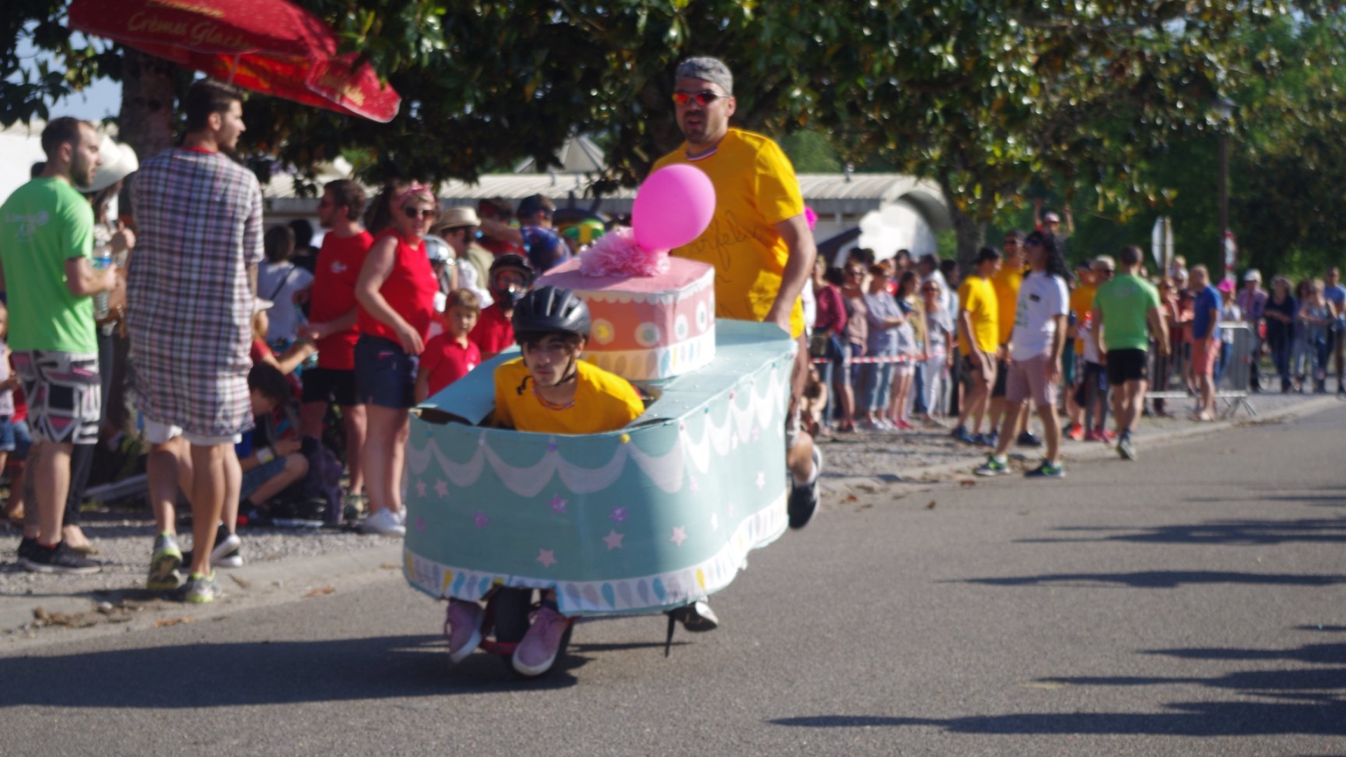 Two men: one in a wheelbarrow, the other pushing it, with a crowd watching the decorated wheelbarrow race at the Fête de l’Andouillette in Mios