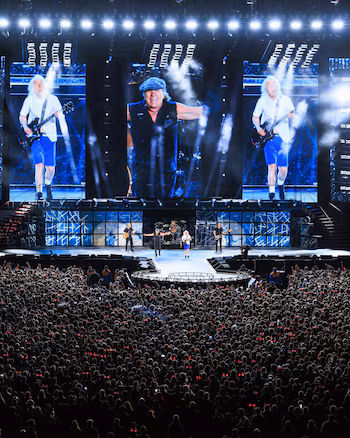 AC/DC concert live: aerial view of a large stage with LED screens showing the musicians and a vast crowd of fans with raised arms