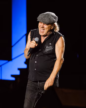 Vocalist Brian Johnson of AC/DC sings passionately into a microphone, wearing a gray cap, black tank top and jeans, on a dark stage with blue lights