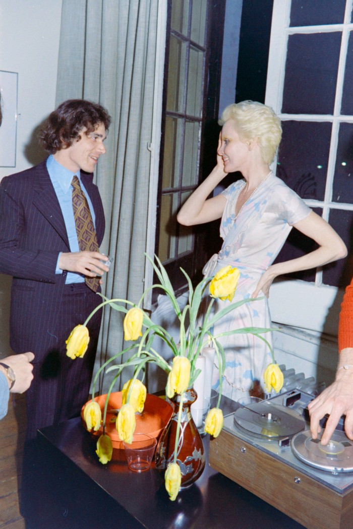 Un hombre con traje sostiene una bebida y habla con una mujer en un estudio tipo loft, con un jarrón de flores sobre una mesa frente a ellos.