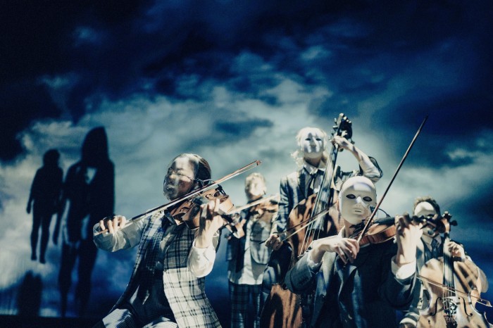 Miembros enmascarados de una orquesta tocan violines y contrabajos sobre un fondo que parece un cielo oscuro y tormentoso.