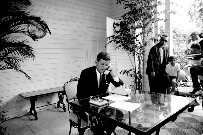 Una fotografía en blanco y negro de un hombre con traje sentado en una mesa con superficie de cristal, hablando por un teléfono de disco.  La habitación presenta plantas en macetas y un grupo de personas, incluido un niño, al fondo.