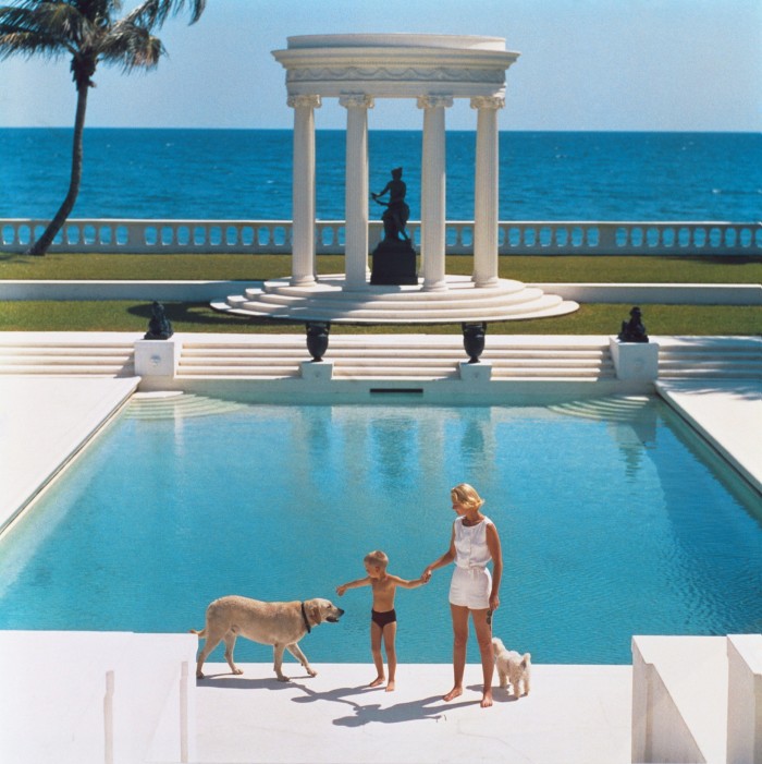Una mujer y un niño caminan de la mano junto a una piscina, acompañados por dos perros.  El telón de fondo presenta una gran columnata blanca y el océano.