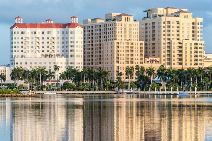 Una vista pintoresca de una ciudad costera con edificios altos de colores claros y exuberantes palmeras a lo largo del paseo marítimo.  Los edificios y los árboles se reflejan en el agua tranquila.