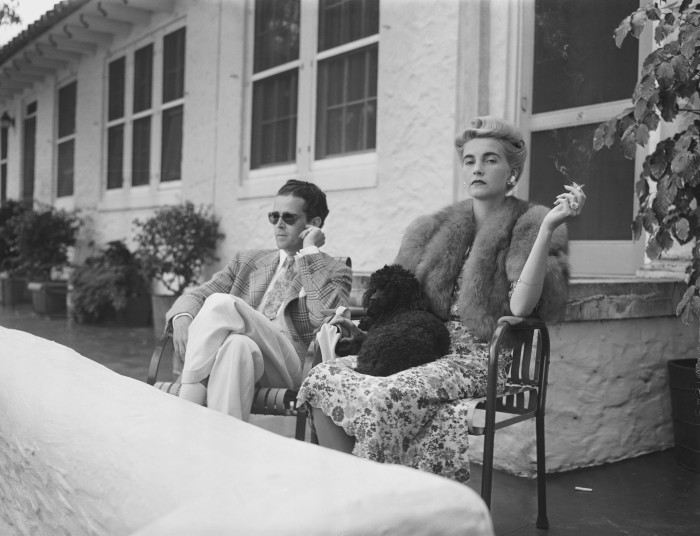 Una fotografía en blanco y negro de una pareja elegante sentada afuera de una casa.  La mujer, vestida con una estola de piel y un vestido de flores, sostiene un cigarrillo y tiene un caniche en el regazo.  El hombre, con gafas de sol y traje a cuadros, está sentado relajado con las piernas cruzadas.