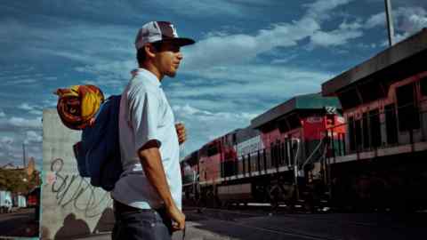 Un hombre con sombrero, una bolsa y una manta a la espalda observa el paso de un tren de mercancías pesadas
