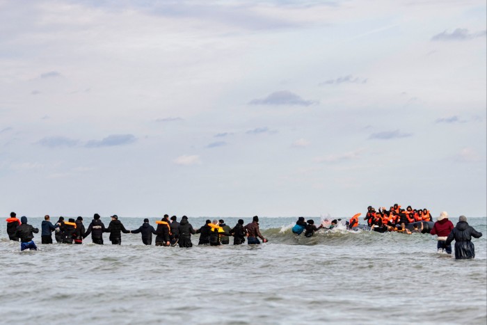 Mucha gente en un pequeño bote de goma con chalecos salvavidas de color naranja.  Hay una fila de personas cogidas de la mano desde la orilla hasta el barco.  Algunas personas luchan en las olas.