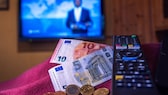 La suma de 18,94 euros está sobre una mesa junto al mando a distancia de un televisor.  Según cálculos preliminares de expertos financieros, el canon de retransmisión aumentará a 18,94 euros a partir de 2025.  Según información de la Agencia de Prensa Alemana, así se desprende de un borrador del comité de expertos del KEF conocido el viernes.