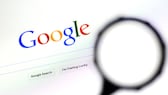 Imagen simbólica: pantalla de inicio de la Búsqueda de Google con una lupa en primer plano