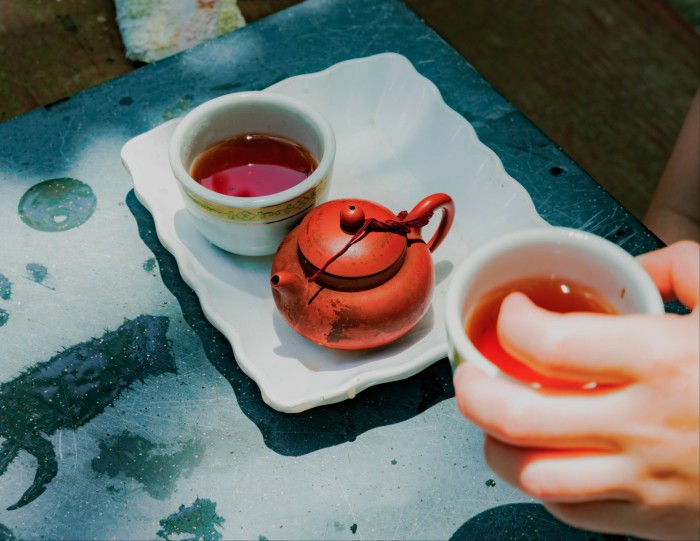 Primer plano de una preparación tradicional para el té. La escena muestra una pequeña tetera roja, una taza de té de porcelana blanca con té y otra mano que sostiene una taza de té llena de té.