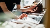 Imagen simbólica: Mujer sosteniendo un teléfono inteligente y escribiendo algo en la computadora portátil