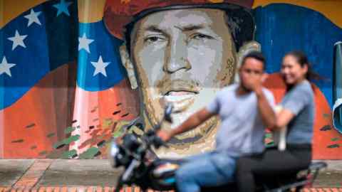 Personas en una motocicleta pasan frente a un mural de Hugo Chávez