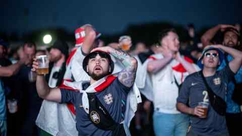 Jóvenes con camisetas de fútbol azules o blancas, algunos de ellos envueltos en banderas de Inglaterra, miran hacia arriba tensos. Algunos de ellos se llevan una mano a la cabeza.