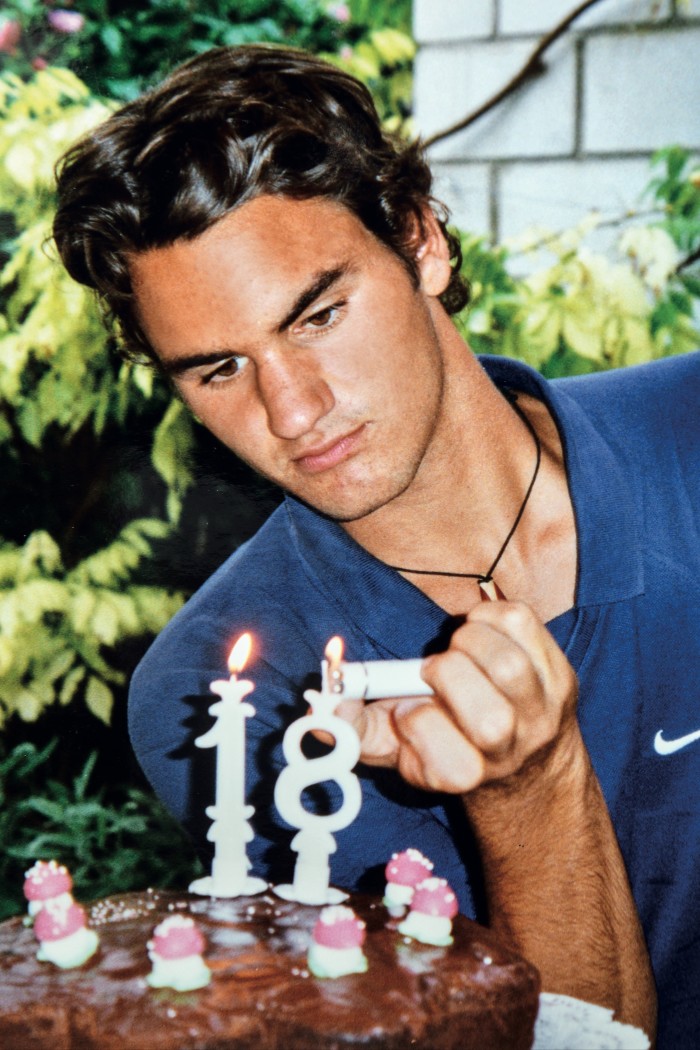 Un joven de cabello oscuro y bronceado, Roger Federer, celebra su cumpleaños número 18 encendiendo velas sobre un gran pastel de chocolate.
