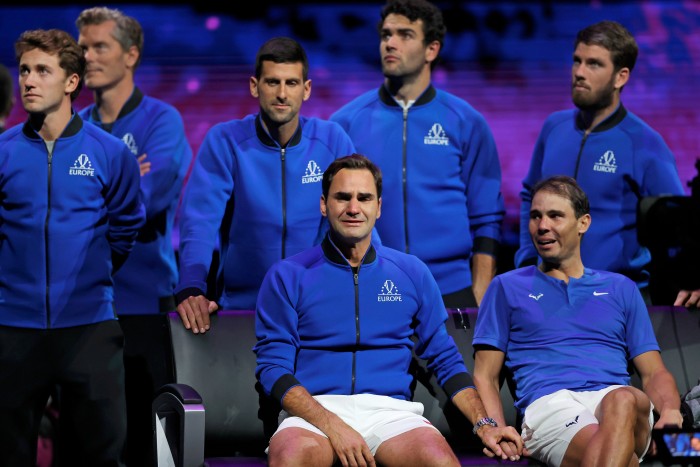 Roger Federer está sentado con su famoso rival Rafael Nadal de la mano y llorando. La pareja, que lleva pantalones cortos blancos y camisetas azules, acaba de perder un partido de dobles en Londres, que fue el último partido de Federer antes de retirarse. Detrás de ellos hay otros jugadores.   