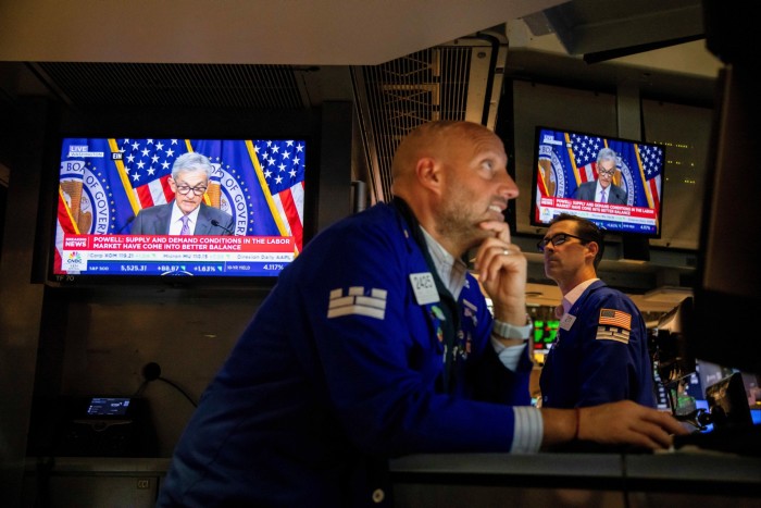 Un operador en la Bolsa de Valores de Nueva York en el trabajo mientras una pantalla de televisión muestra al jefe de la Reserva Federal, Jay Powell, hablando