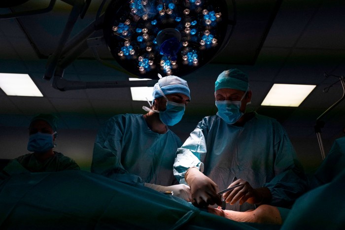 Los cirujanos realizan una operación a un paciente bajo la luz brillante de un quirófano en el centro Georges-Francois Leclerc en Dijon, Francia. Están administrando quimioterapia intraperitoneal en aerosol presurizada (PIPAC) para tratar las metástasis peritoneales del cáncer de páncreas y de las vías biliares. El equipo quirúrgico está vestido con batas quirúrgicas azules, mascarillas y gorros.