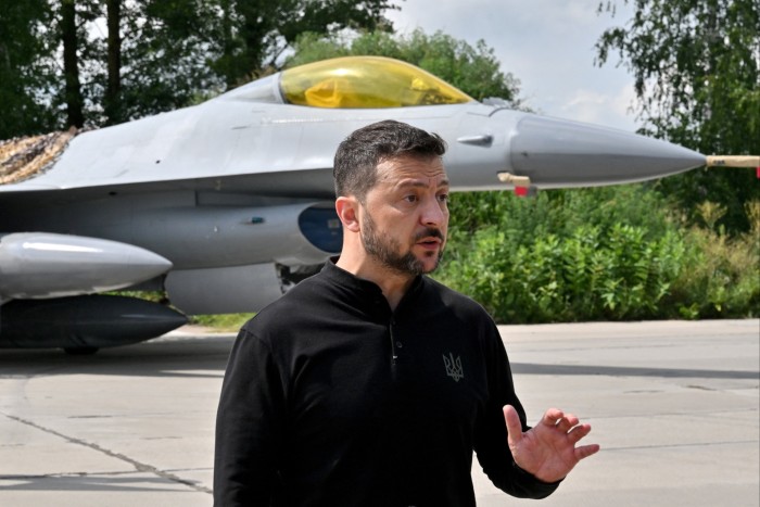 Volodymyr Zelenskyy hace un gesto mientras habla con representantes de los medios de comunicación mientras está de pie frente a un avión de combate F16.