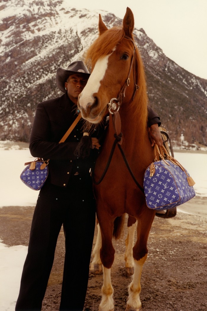 Un modelo con pantalones negros, chaqueta y sombrero de vaquero está de pie con el brazo sobre el cuello de un caballo. Detrás de ellos hay una montaña cubierta de nieve. 