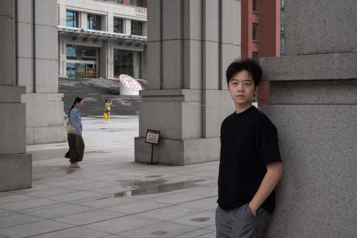 Rui Pang, estudiante de grado del Programa de Gestión Internacional ACE de la Universidad Renmin de Pekín, posa para un retrato apoyado en una columna. Al fondo, pasa otra persona y una tercera se toma una foto cerca de la entrada del edificio.