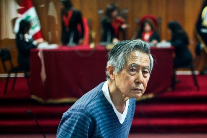 Alberto Fujimori, expresidente de Perú, es visto en una sala de tribunal durante una audiencia en Lima el 7 de noviembre de 2013. Al fondo, varias personas, incluidos jueces, están sentadas en una mesa con un mantel rojo y la bandera peruana visible.