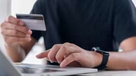 Un hombre sentado frente a su computadora portátil sostiene una tarjeta de crédito en la mano. 