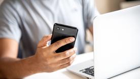 Un hombre se sienta frente a una computadora portátil y sostiene un teléfono inteligente negro en su mano derecha.