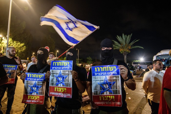 Los manifestantes enmascarados sostienen pancartas. Al fondo ondea una bandera israelí.