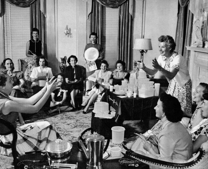 Brownie Wise muestra con entusiasmo un recipiente Tupperware lleno de agua a un grupo de mujeres en una fiesta Tupperware en la década de 1950. Las mujeres están reunidas en una sala de estar, sentadas alrededor de una mesa que muestra varios productos Tupperware.