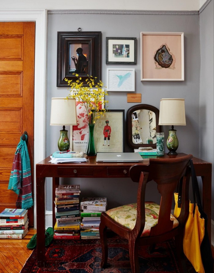 Una habitación con un escritorio y una mesa en la que hay un ordenador portátil, fotografías y dos lámparas, con cuadros en la pared y pilas de libros en el suelo.