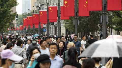 Una gran multitud camina por la calle Nanjing East Road en Shanghái, China. La calle está decorada con numerosas banderas nacionales chinas y la zona está rodeada de árboles y varias tiendas.