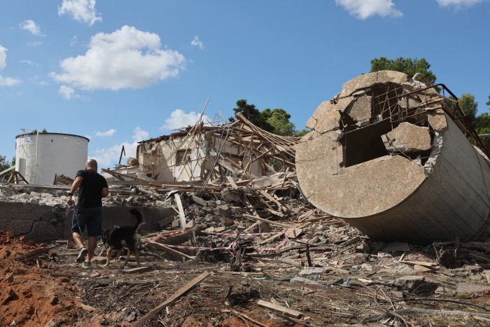 Un hombre camina con un perro entre los escombros de un edificio destruido. Los escombros y las estructuras dañadas son visibles bajo un cielo parcialmente nublado.