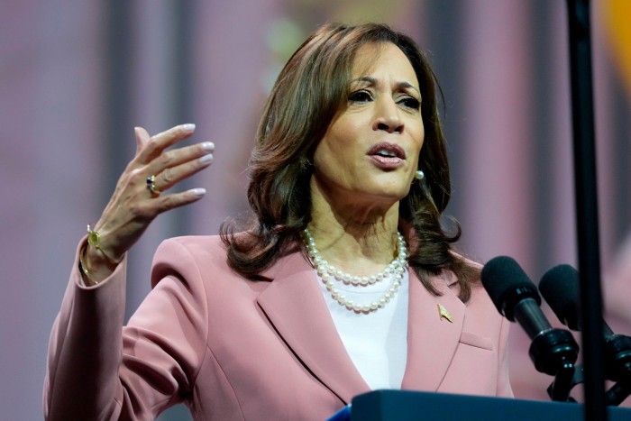 Kamala Harris habla en un podio y hace un gesto con una mano. Lleva una chaqueta rosa sobre un top blanco.