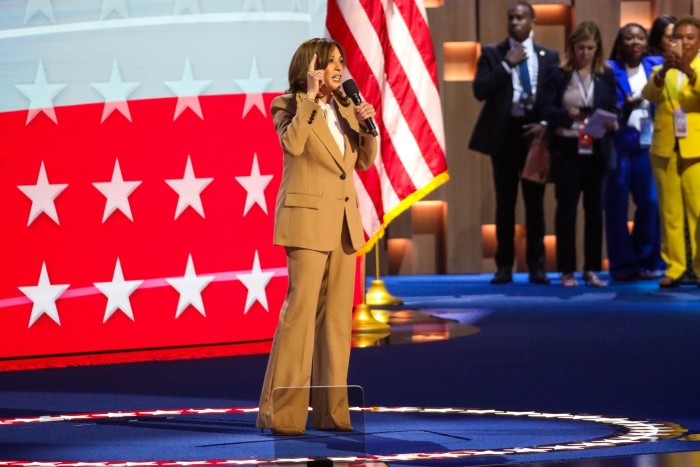 Kamala Harris está hablando en un escenario, con el micrófono en una mano y la otra apuntando hacia arriba. Detrás de ella hay una enorme bandera de barras y estrellas.