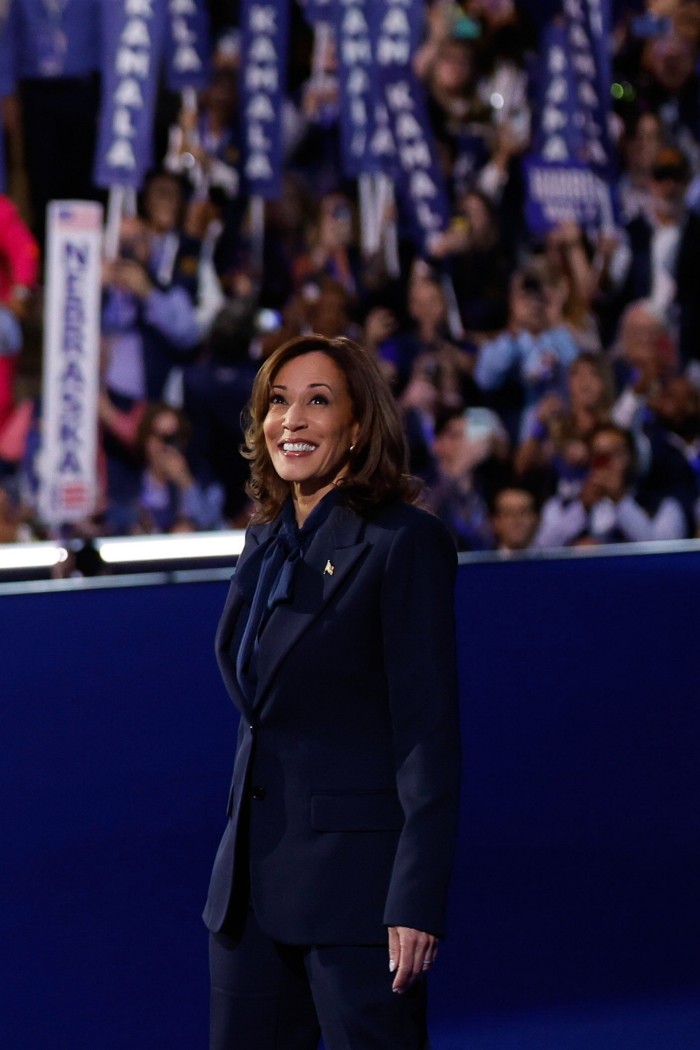 Kamala Harris sonríe mientras una multitud de personas detrás de ella sostiene pancartas que dicen su nombre. Lleva un traje oscuro y una blusa oscura con lazo.