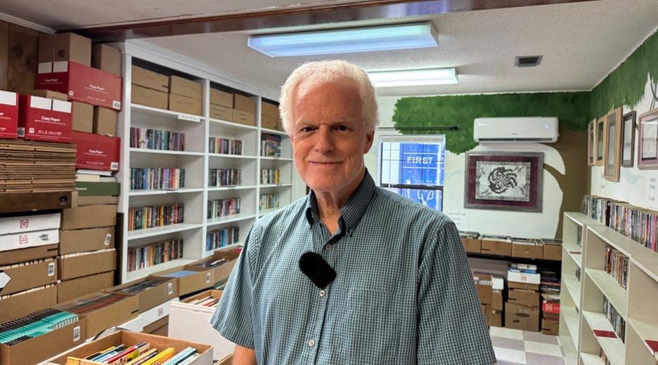 Un hombre con una camisa corta a cuadros se encuentra en una habitación llena de libros y mira amigablemente a la cámara.