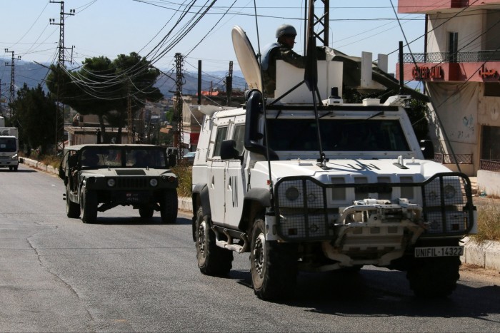 Un vehículo de mantenimiento de la paz de la ONU, marcado con "Naciones Unidas" y una matrícula de Unifil, conduce junto a un vehículo del ejército libanés en el sur del Líbano. En la torreta del vehículo de la ONU se ve a un soldado. Al fondo se ven edificios y líneas eléctricas.