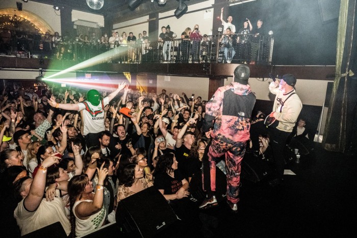 Vista desde detrás de dos hombres con ropa deportiva y gorras de béisbol cantando en el escenario en una sala repleta de un público que saludaba con las manos