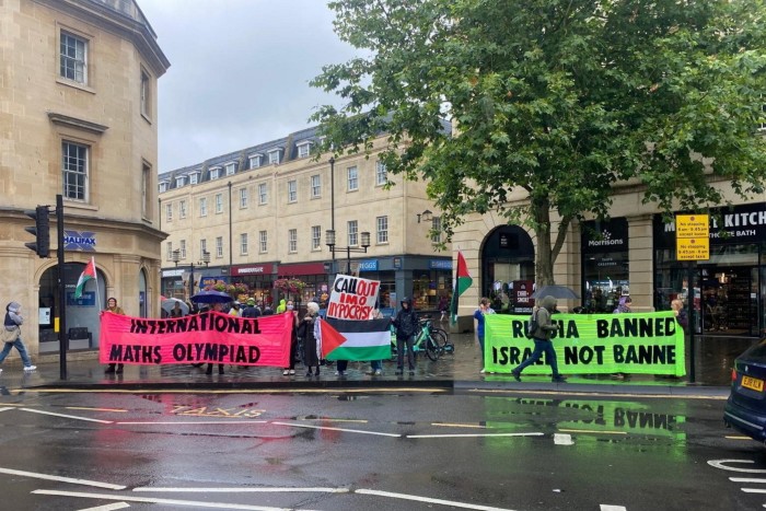 Los manifestantes están en la calle sosteniendo pancartas durante la final de la OMI. Una pancarta rosa dice “Olimpiada Internacional de Matemáticas”, mientras que otra dice “Llame a la hipocresía de la OMI” con un diseño de bandera palestina. Una pancarta verde dice “Rusia prohibida, Israel no”. 