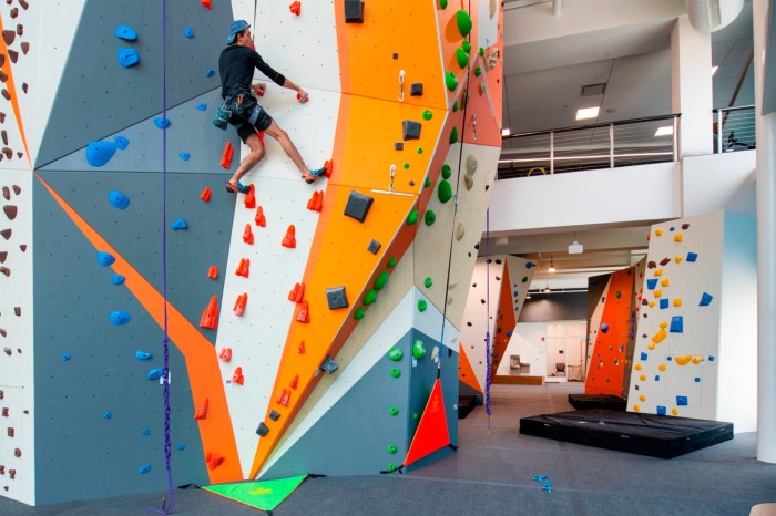 Un gimnasio de escalada cubierto con vibrantes muros de escalada multicolores. Un escalador con arnés y equipo de seguridad está a mitad de un ascenso.