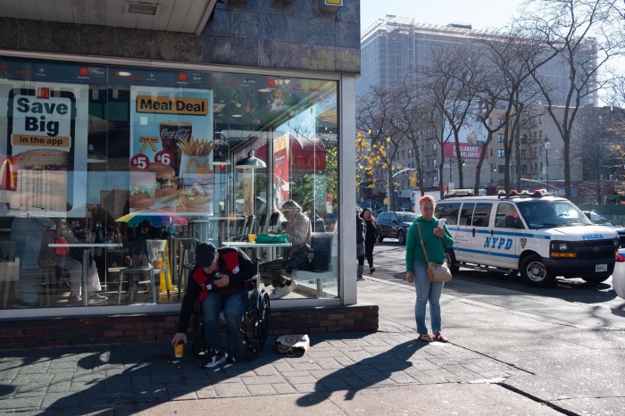 Vista de la calle del Bronx que muestra a peatones, un hombre sentado en la acera frente a un restaurante de comida rápida y un coche de policía esperando en el tráfico.