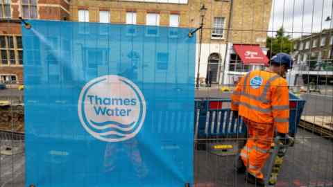 Un cartel con el logotipo de Thames Water está pegado a las barreras protectoras en un lugar de trabajo de suministro de agua en Londres. Al fondo, dentro del área vallada, se ve a un trabajador con un uniforme naranja de alta visibilidad.