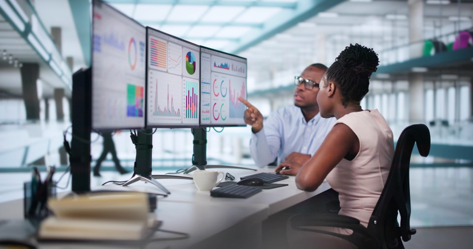 Dos analistas de negocios que revisan datos en una configuración de monitor triple.
