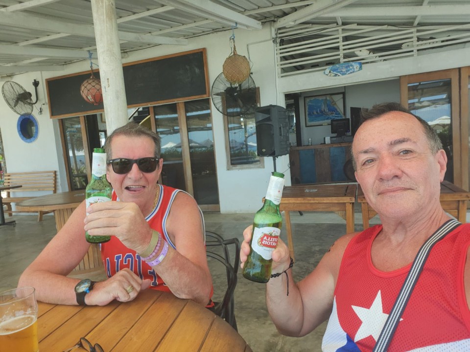 Dos hombres sentados en una mesa al aire libre, cada uno sosteniendo una botella de cerveza.