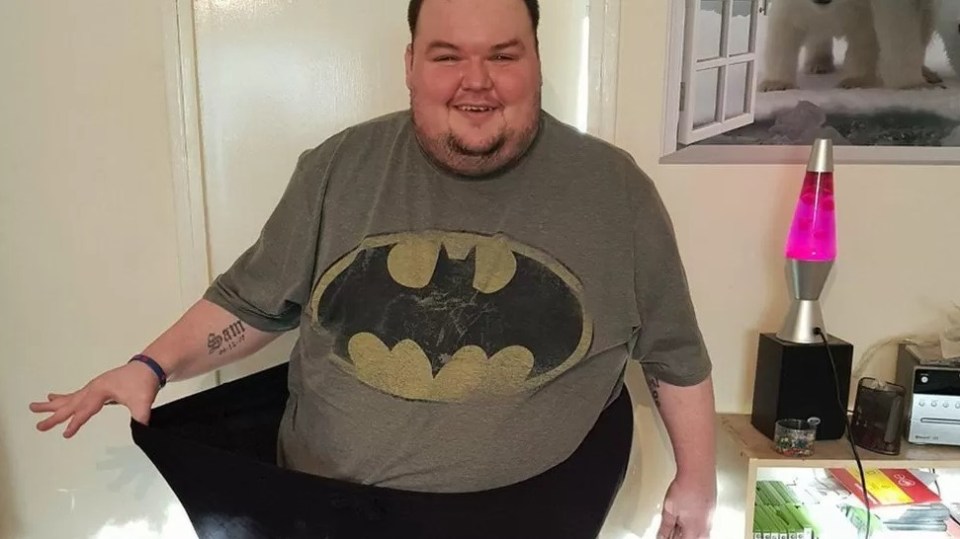 El hombre con la camisa de Batman sosteniendo pantalones grandes para mostrar la pérdida de peso.