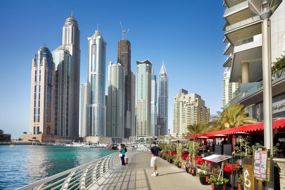 Dubai Marina Waterfront con rascacielos y personas caminando por un paseo marítimo con restaurantes.
