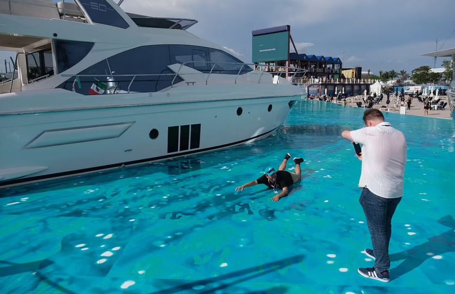 Una persona se encuentra en una superficie de agua falsa junto a un yate mientras otra persona toma una foto.