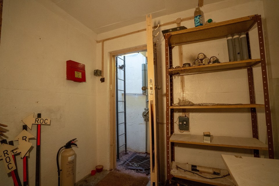 Interior de un búnker de la Guerra Fría, que muestra una puerta con una escalera que conduce hacia abajo y los estantes con varios artículos.