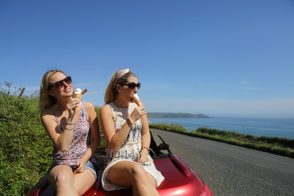 Dos mujeres comiendo conos de helado mientras están sentadas en un convertible rojo con vistas al océano.
