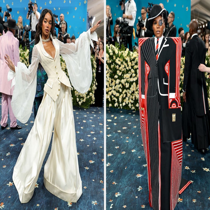 Dos individuos en una alfombra roja: una con atuendo blanco que fluye, el otro con un atuendo abstracto llamativo con chaqueta y sombrero de hombros anchos